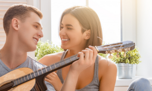 young man plays guitar happily smiles at girlfriend sitting next to