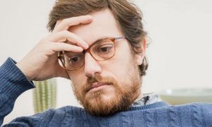 man hand on forehead wistful eyes sits in living room worrying thinking about his emotions