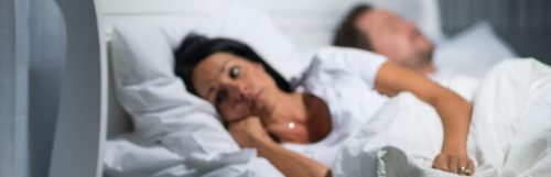 middle aged woman lying awake on bed while husband sleeping nicely