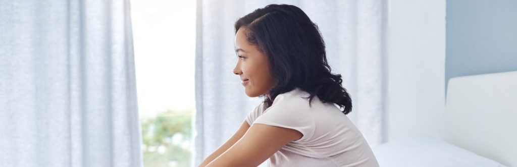 young woman sitting on bed looking through window happily smiling