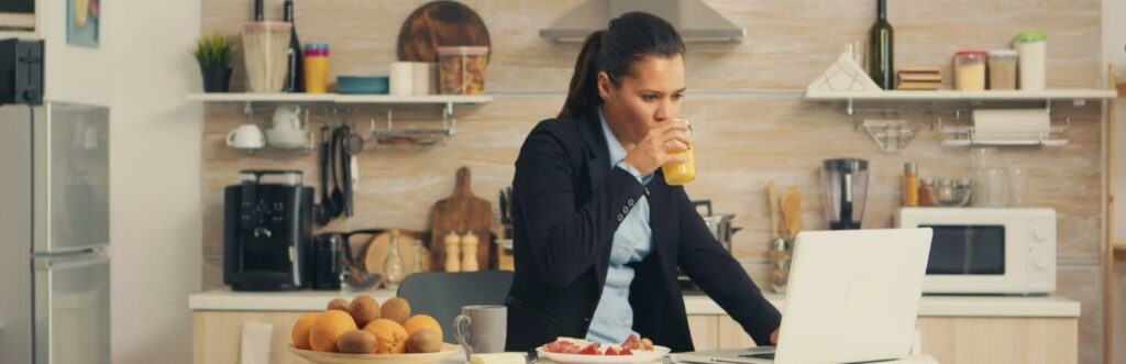 business woman stands in kitchen beside fruit dish drinks orange juice looks at laptop