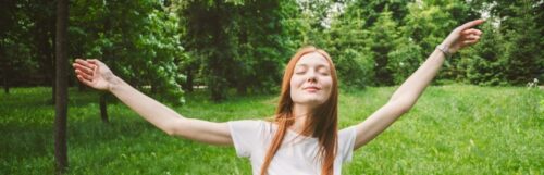 young woman raises hands eyes closed happily enjoys immerses into nature stands in forest