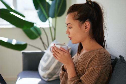 young woman sits on dark blue couch holding white tea mug in quiet living room