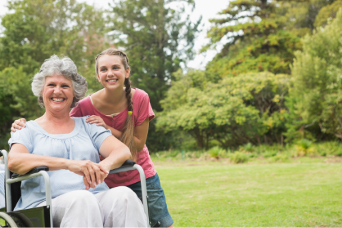 maternal grandmother sits on wheelchair smiling besides granddaughter standing smiling in park 