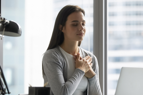 Peaceful young woman worker sit at workplace hold hands at heart chest feel grateful thankful for good luck or fate, millennial female employee believer meditate pray at desk in office, faith concept
