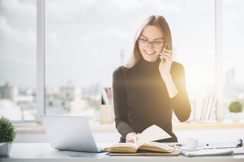 Happy smiling european businesswoman sitting at office desk, talking on cellphone, working on project and doing paperwork. Blurry city view with sunlight in the background