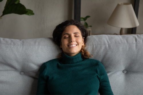 Happy millennial Caucasian woman sit rest on cozy couch in living room relieve negative emotions. Smiling young female relax on sofa at home with eyes closed, sleep or daydream on weekend.