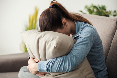 Unhappy lonely depressed woman at home, she is sitting on the couch and hiding her face on a pillow, depression concept