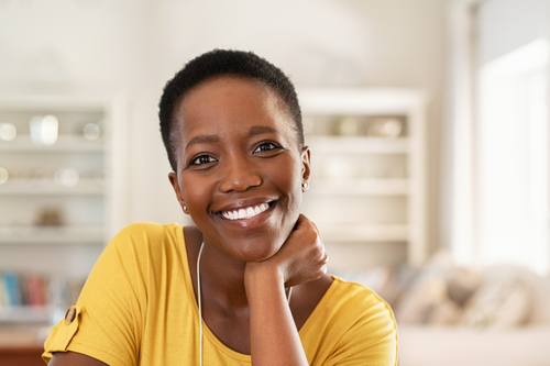 Portrait of middle-aged Black woman sitting at home and looking at camera. Cheerful black girl with short hair in casual sitting in her new apartment with copy space. Satisfied woman smiling while looking at camera.