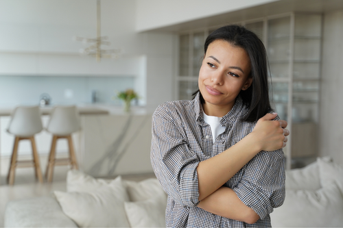 Happy serene woman gently embraces herself, expresses self love feels comfortable, standing at cozy home. Calm female hugs herself and smiling with pleasure, feeling self-pride, dreaming.