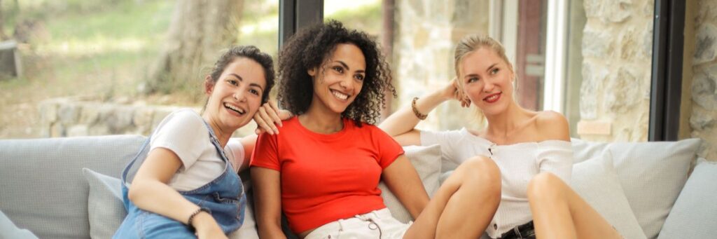 Three friends laughing together on a bright sofa, collective energy at its best
