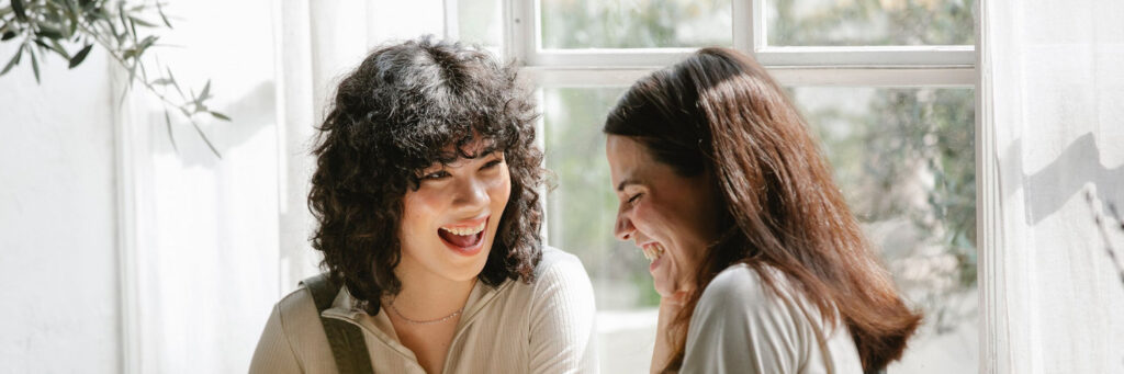 Two women in their forties laughing together in a bright, light-filled room, showing emotional resonance at its best