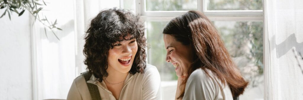 Two women in their forties laughing together in a bright, light-filled room, showing emotional resonance at its best