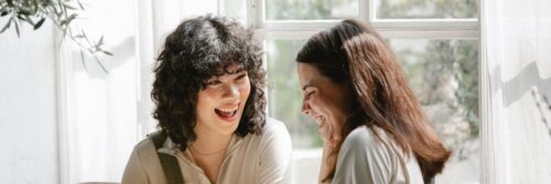 Two women in their forties laughing together in a bright, light-filled room, showing emotional resonance at its best