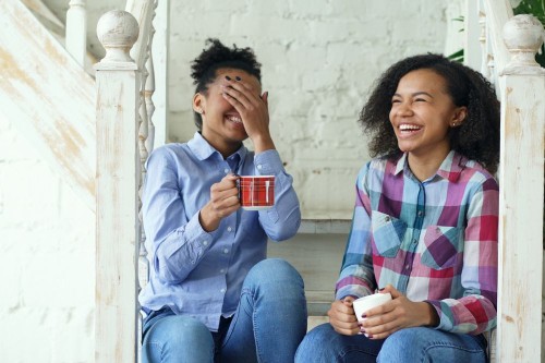 Two friends laughing together on the stairs with mugs of tea, relaxed in a secure friendship