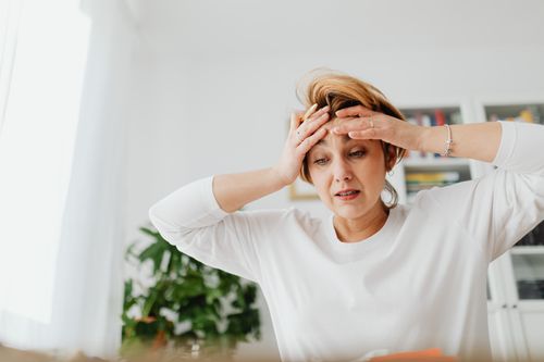 A tense woman sitting on a bright modern sofa rubbing her neck, showing what life feels like without nervous system regulation