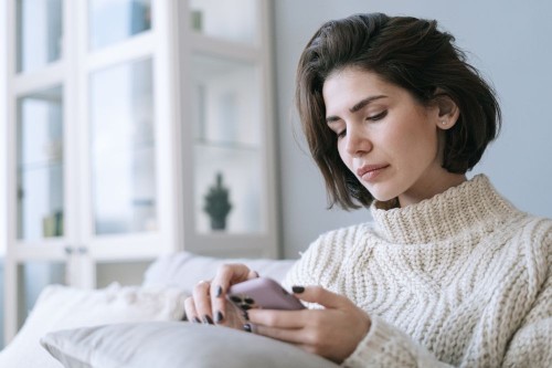 A woman on a sofa anxiously watching her phone for a reply, a common sign of fear of abandonment