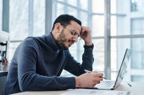 A man in glasses resting his head in his hand at his laptop in a bright office, a sign of poorly managed job demands at work