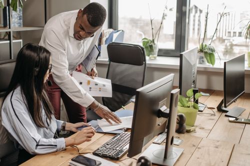 A manager leaning in to talk with a colleague at her desk in an open-plan office, beginning the work of managing job demands at work