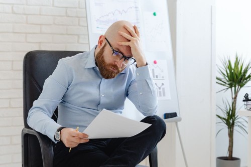 A worker holding his head as he reads a demanding report, a sign of high job demands, one of the most common psychosocial hazards at work