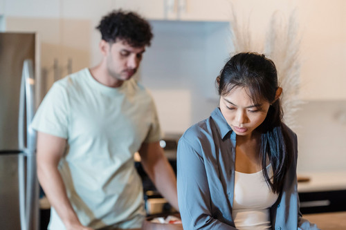 A woman sitting apart from her partner with a tense expression in a bright living room, the daily reality of trust issues in relationships