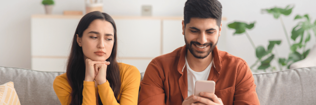 Trust issues in relationships between a woman with dark hair in a yellow top and a man in a brown shirt looking at his phone