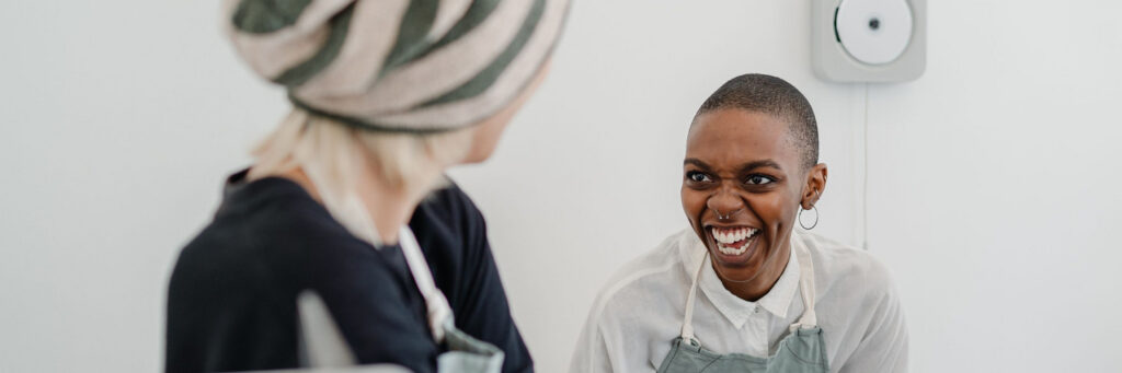 Two women in their forties laughing together in a bright modern kitchen, showing life without chronic stress symptoms