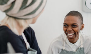 Two women in their forties laughing together in a bright modern kitchen, showing life without chronic stress symptoms