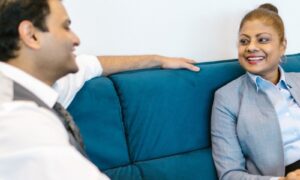 Two colleagues in their forties sitting on a blue sofa in easy conversation over coffee, listening to each other rather than people pleasing
