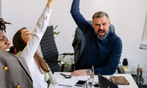 A manager and two colleagues laughing together around a table in a bright modern office, showing what happens after learning how to review workload at work