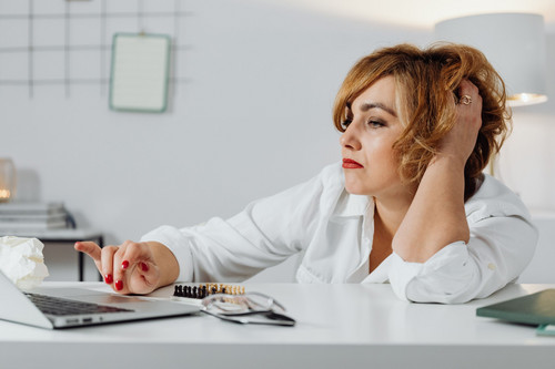 A woman sitting at a laptop looking tense and uncertain in a bright office, showing what imposter syndrome feels like