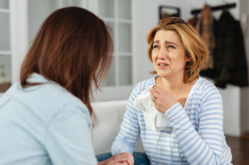 A woman looking drained while listening to a friend talking in a bright room, showing emotional contagion taking a toll
