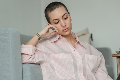 A woman in her forties sitting quietly by a window in a bright apartment, looking flat and drained by low energy emotions