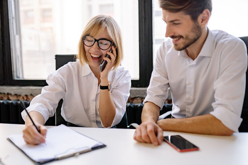 Two colleagues laughing together at a desk in a bright office, showing a hybrid team at ease after remote work psychosocial risks are managed
