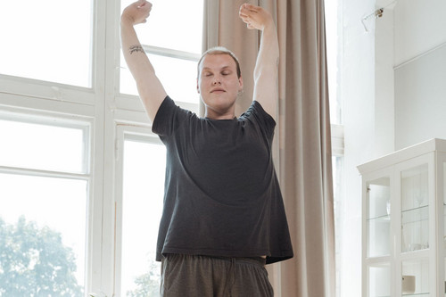 A man standing and stretching beside a sunlit window at home, the first small step in rebuilding movement and energy levels