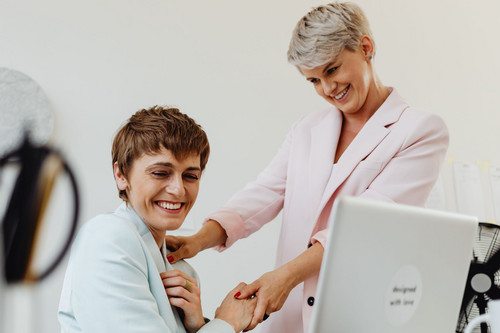 Two colleagues in their forties talking warmly across a desk in a bright office, the kind of honest conversation that eases imposter syndrome