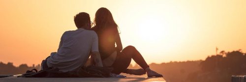 couple sits on rock facing backward watching sunset