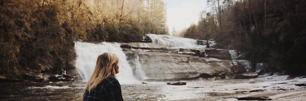 woman stands beside waterfall looking at forest in sunny sky