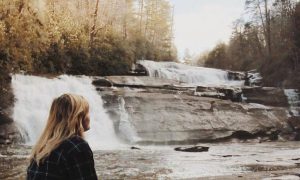 woman stands beside waterfall looking at forest in sunny sky