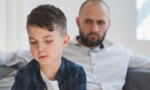 son feels sad while father sitting on couch behind angrily looking at him