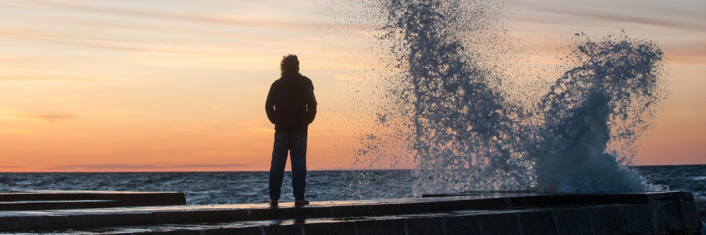 man stands on seaport looking at strong ocean wave in sunset sky