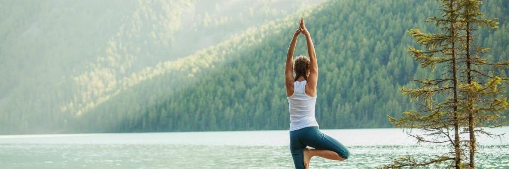 woman stands practicing yoga near lake in beautiful sunny sky