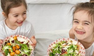 two little adorable girls happily smile sits on bed holding two healthy vegetable dishes