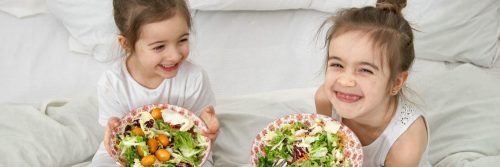 two little adorable girls happily smile sits on bed holding two healthy vegetable dishes