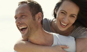 man excitedly carries girlfriend on back while woman happily laughing in cloudy sky