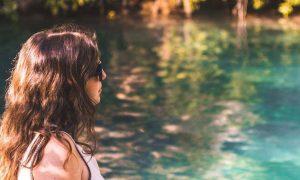 woman wearing sunglasses stands beside blue water lake thinking