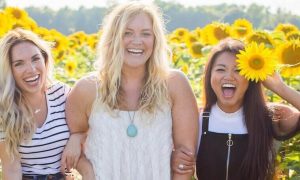 three women stand within sunflower garden happily smiling posing for photos