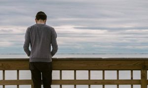 man facing backward stands alone on bridge looking at ocean in cloudy sky