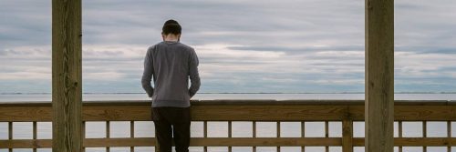 man facing backward stands alone on bridge looking at ocean in cloudy sky