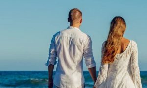 couple stands facing backward holding hands in front of blue ocean in blue clear sky
